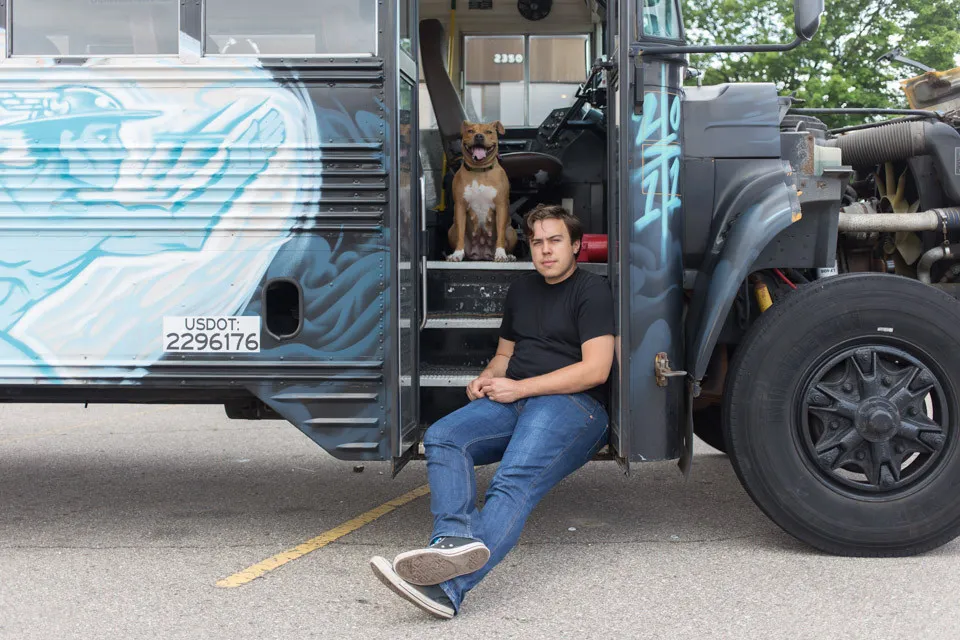 Andy Didorosi, founder of The Detroit Bus Company, sitting on the step of a hand-painted bus with a dog looking on