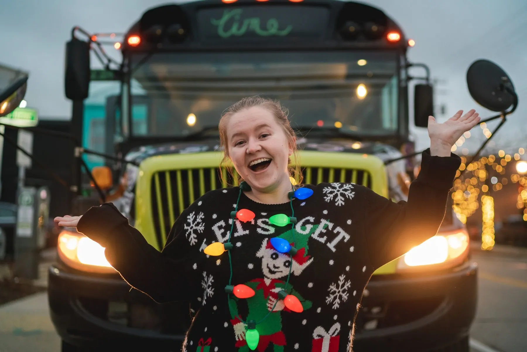 A grinning guest in an ugly sweater, arms out, in front of the bus grille and glowing headlights
