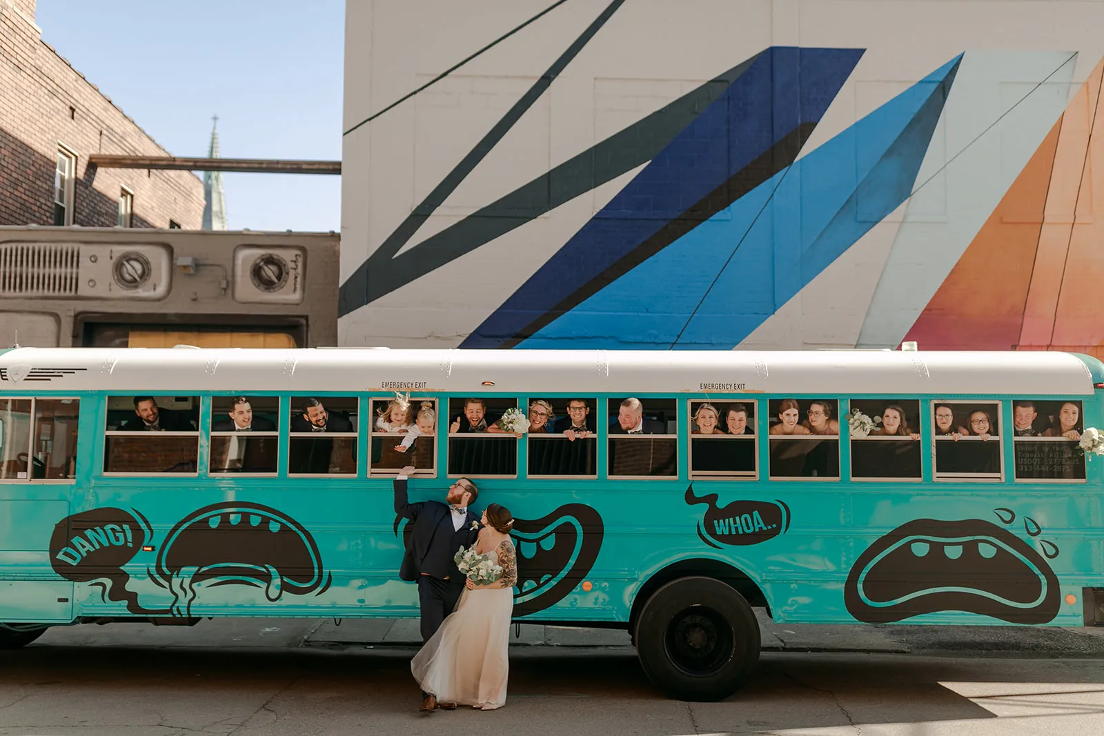 A newlywed couple in front of a teal hand-painted bus beneath a mural